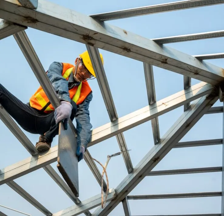Handwerker mit Helm und Warnweste arbeitet auf einem Stahldachstuhl vor blauem Himmel